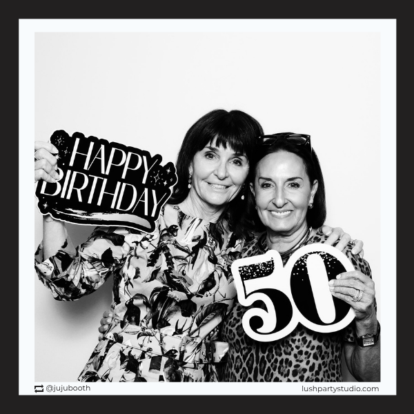 Two women holding 'Happy Birthday' and '50' prop signs in a photo booth setting.