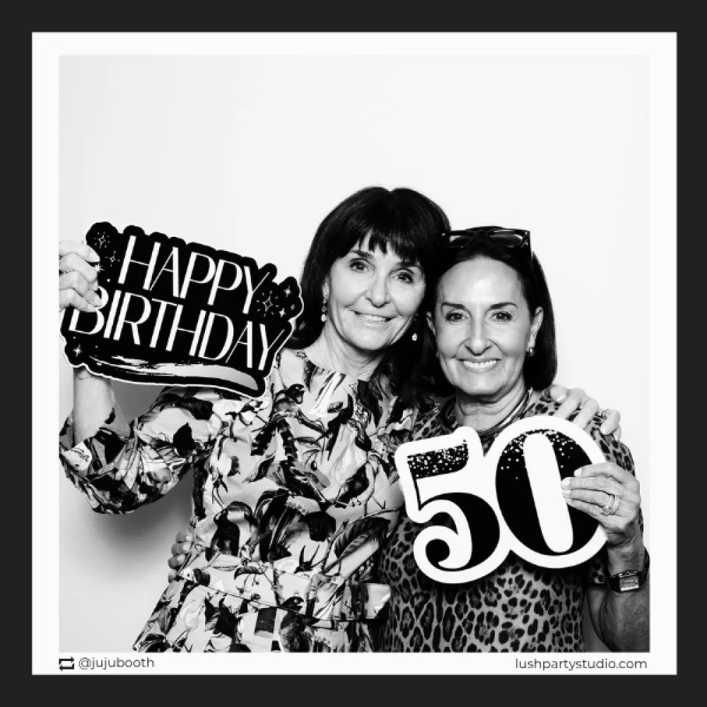 Two women holding 'Happy Birthday' and '50' prop signs in a photo booth setting.