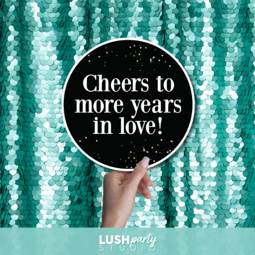 Hand holding a sign with 'Cheers to more years in love!' text by Lush Party Studio.