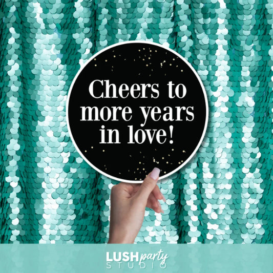 Hand holding a sign with 'Cheers to more years in love!' text by Lush Party Studio.