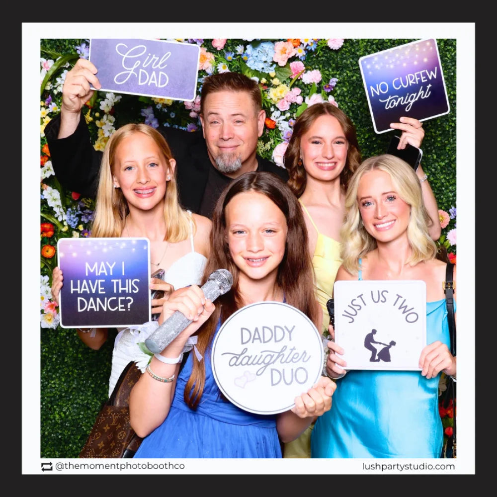 Group of people holding prop signs with daddy daughter dance phrases, posing against a floral backdrop.