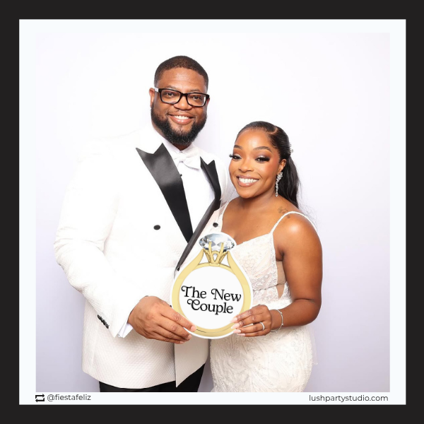 Man and woman in formal attire holding a sign with 'The New Couple' and a large diamond ring prop.