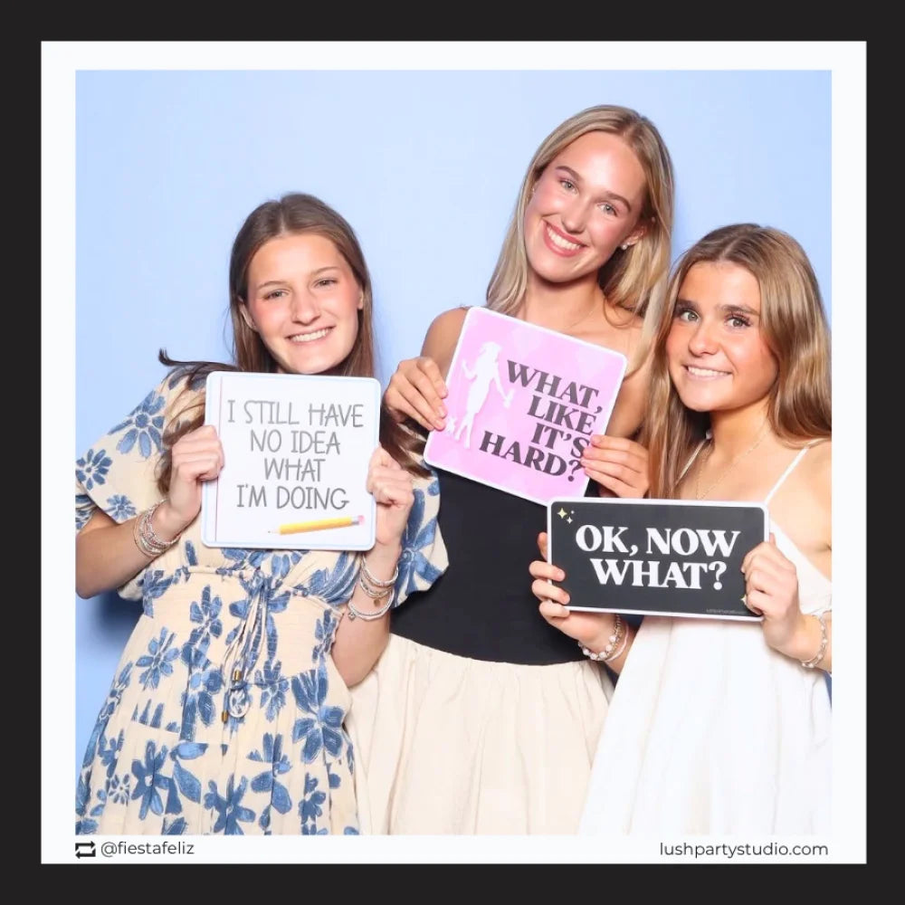 Three women holding photo booth prop signs with graduation phrases in a photo booth setting.