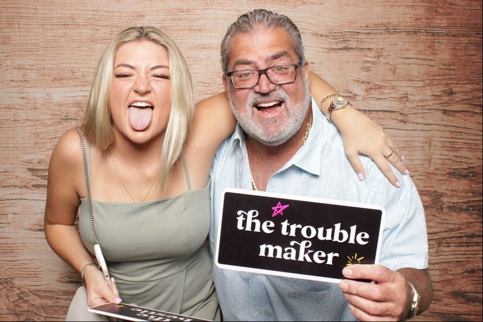 Two people posing with a sign that reads 'the trouble maker' against a wooden background.