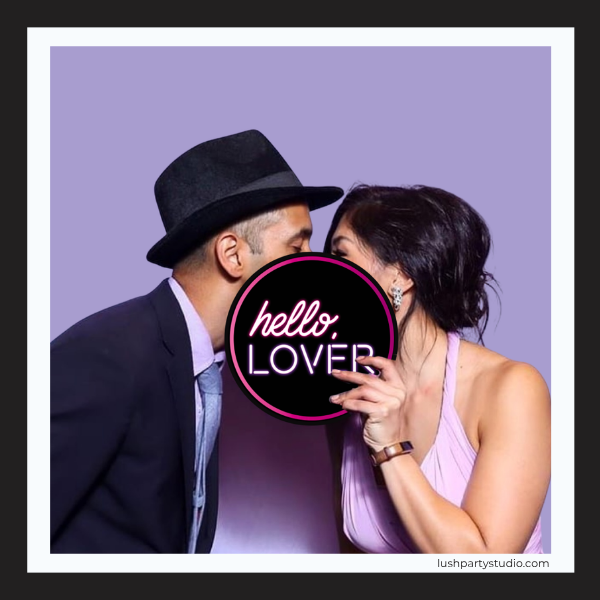 Couple posing with a 'hello, LOVER' photo booth prop sign in neon style
