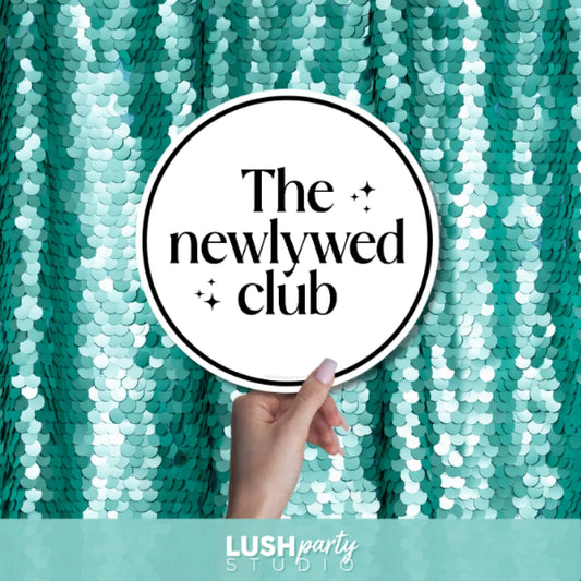 Hand holding a photo prop sign with 'The Newlywed Club' text by Lush Party Studio.