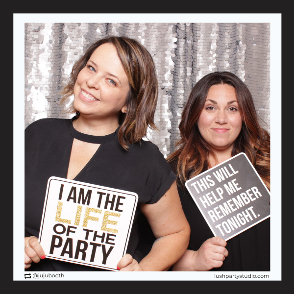 Two women holding photo prop signs with humorous phrases in front of a sparkly backdrop