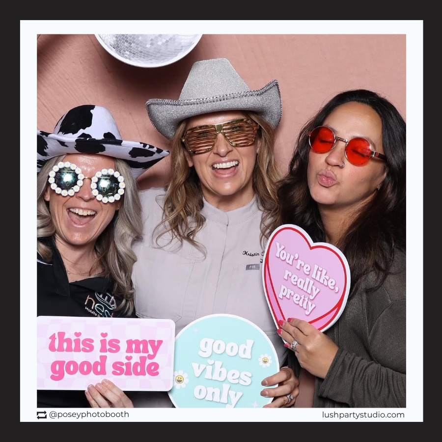 Three people in a photo booth with props and sign by lush party studio