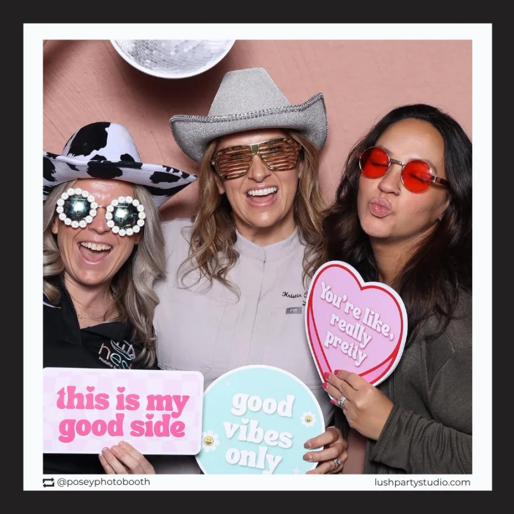 Three people in a photo booth with props and sign by lush party studio