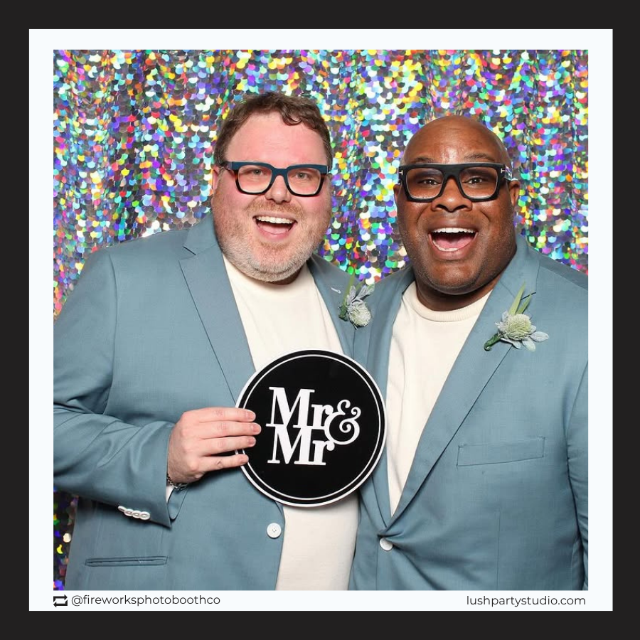 Two grooms holding a 'Mr & Mr' photo booth prop sign for same-sex weddings