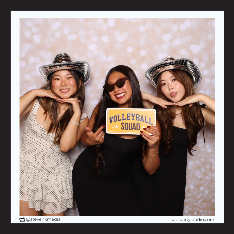 Three women posing with a 'Volleyball Squad' photobooth prop sign in a photo booth with a sparkly background.