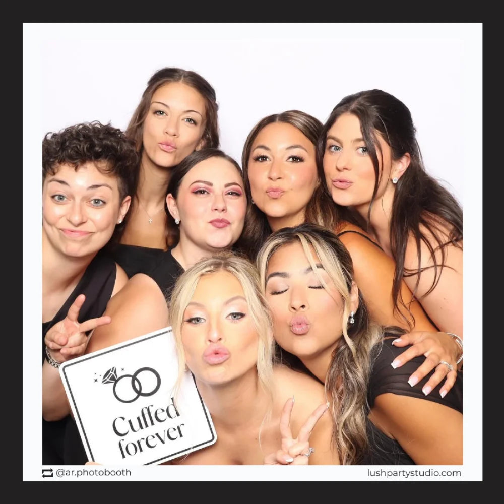 Group of women posing together with a 'Cuffed forever' photo booth prop sign in a photo booth setting by lush party studio. vogue black and white wedding props
