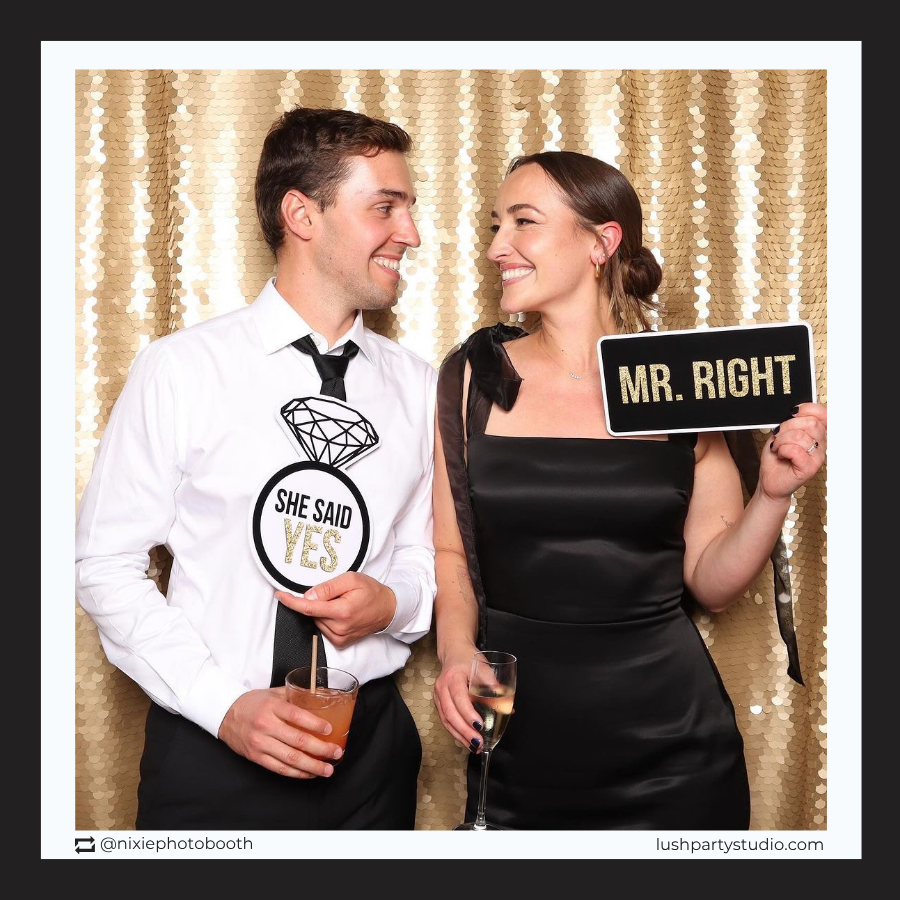 Man and woman posing with 'She Said Yes' and 'Mr. Right' prop signs in a photo booth with a gold sequin backdrop.