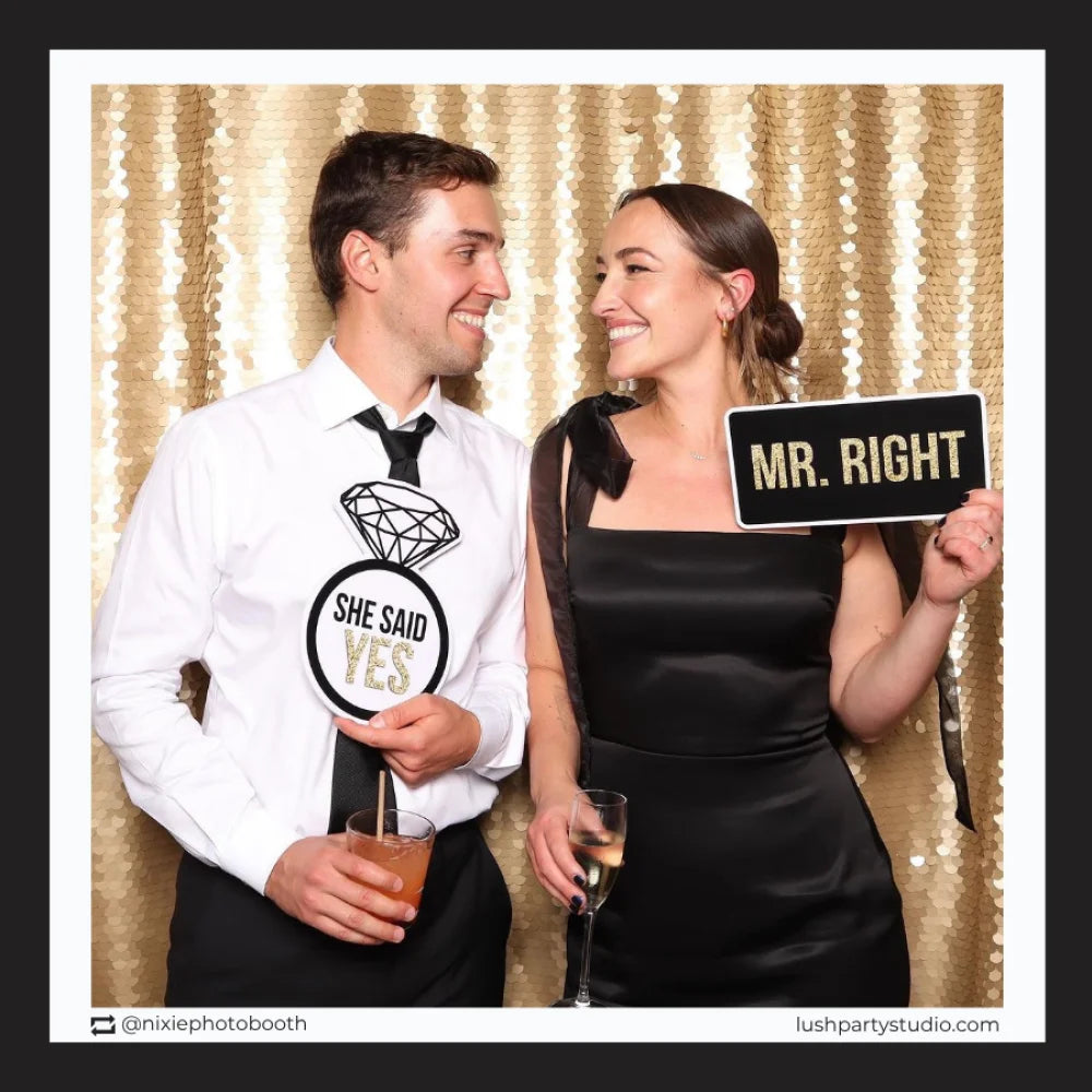 Man and woman posing with 'She Said Yes' and 'Mr. Right' prop signs in a photo booth with a gold sequin backdrop.