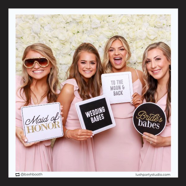 Four women in pink dresses holding photobooth prop signs with wedding-related phrases by lush party studio