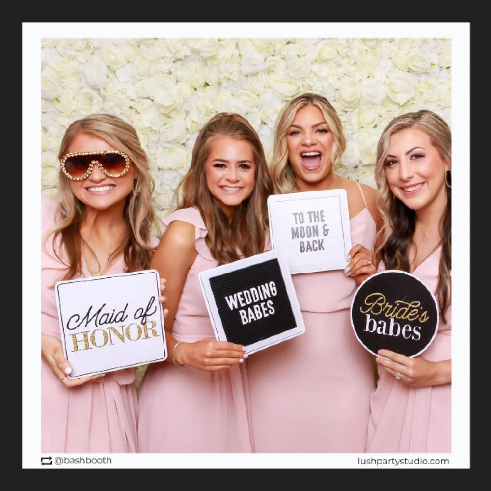 Four women in pink dresses holding photobooth prop signs with wedding-related phrases by lush party studio
