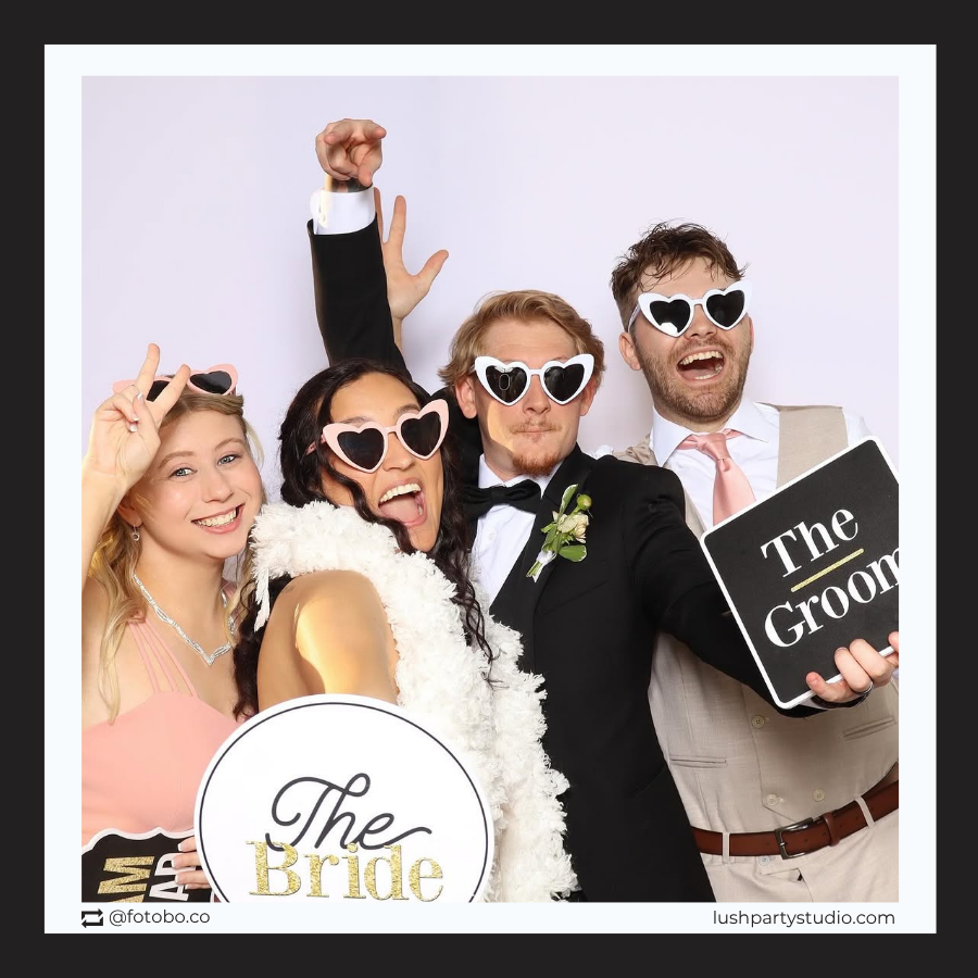 Group of people in formal attire with 'The Bride' and 'The Groom' photo booth prop signs, at a photobooth. 