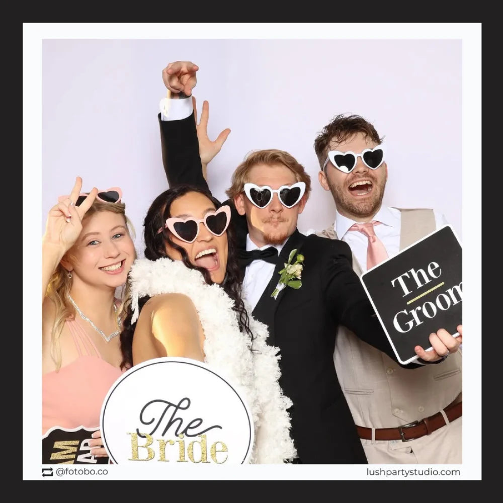Group of people in formal attire with 'The Bride' and 'The Groom' photo booth prop signs, at a photobooth. 