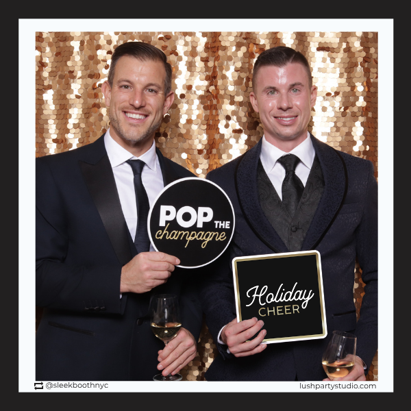 Two men in suits holding 'Pop the Champagne' and 'Holiday Cheer' prop signs